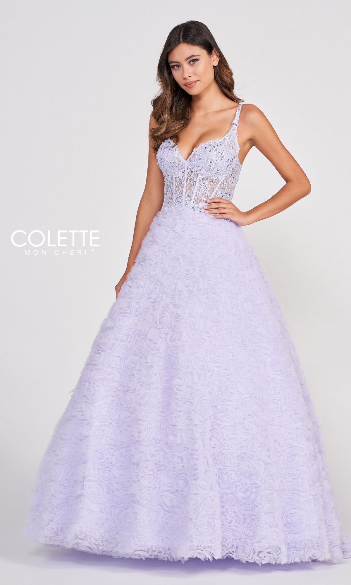 A woman stands against a plain background in the Colette Sheer-Bodice Rosette Beaded Lace Ball Gown, featuring a floor-length, light purple skirt with floral rosettes and a fitted, embellished sheer bodice.